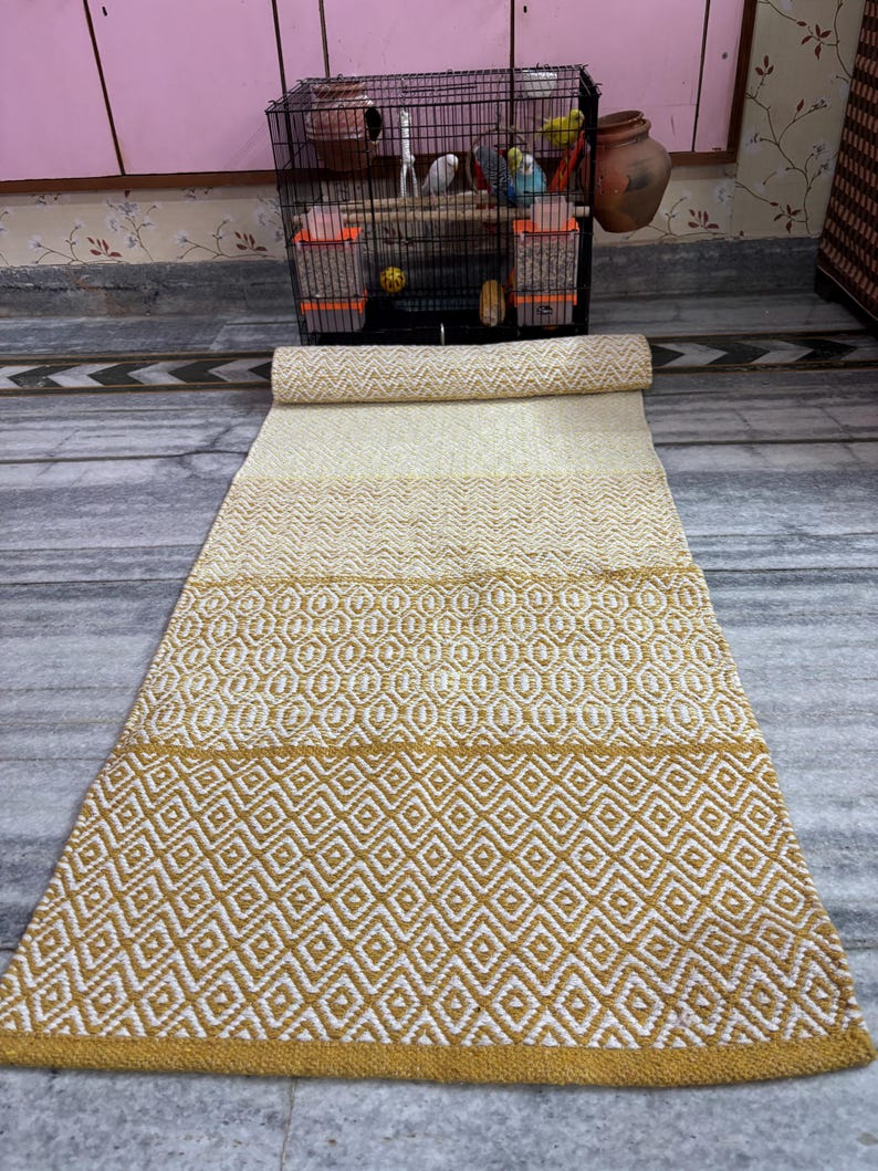 Puede incluir: Una alfombra rectangular enrollada con un patr&oacute;n geom&eacute;trico. La alfombra presenta bandas alternas de crema y amarillo mostaza, con un patr&oacute;n de diamantes en la secci&oacute;n inferior. La alfombra est&aacute; hecha de material tejido.