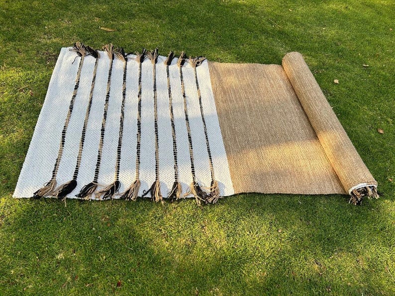 May include: A rectangular, woven rug with a white and black striped pattern on one end and a rolled-up tan section. The rug has decorative tassels on the striped end, suitable for indoor or outdoor use.