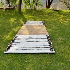 May include: A rectangular rug with a brown center section, transitioning to white with black and brown stripes. The rug has a fringe on both ends and is displayed on a green grassy surface. The rug is designed for indoor or outdoor use.