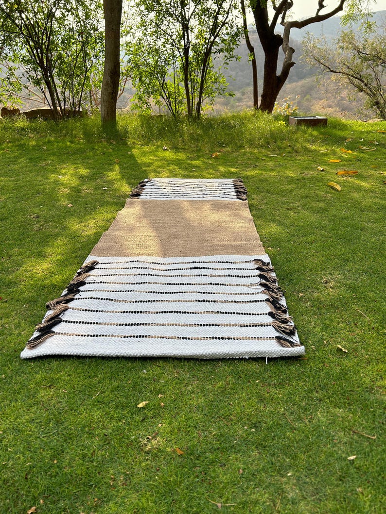 May include: A rectangular rug with a brown center section, transitioning to white with black and brown stripes. The rug has a fringe on both ends and is displayed on a green grassy surface. The rug is designed for indoor or outdoor use.