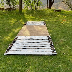 May include: A rectangular rug with a brown center section, transitioning to white with black and brown stripes. The rug has a fringe on both ends and is displayed on a green grassy surface. The rug is designed for indoor or outdoor use.