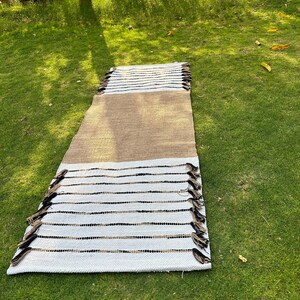 May include: A rectangular rug with a woven design. The rug features a central tan section, bordered by white sections with black and tan stripes. The edges are finished with tassels. The rug is displayed on green grass.