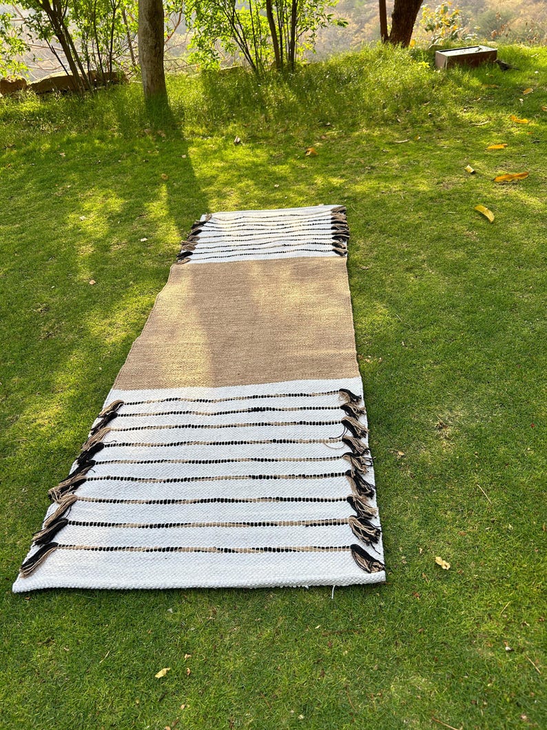 May include: A rectangular rug with a woven design. The rug features a central tan section, bordered by white sections with black and tan stripes. The edges are finished with tassels. The rug is displayed on green grass.
