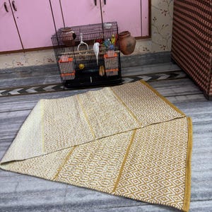 Puede incluir: Una alfombra rectangular plegada con un patr&oacute;n geom&eacute;trico en tonos crema y dorado. La alfombra est&aacute; hecha de material tejido y tiene un borde dorado. Una jaula para p&aacute;jaros con p&aacute;jaros y un armario rosa est&aacute;n en el fondo.