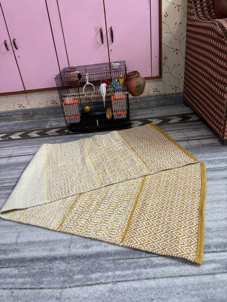 Puede incluir: Una alfombra rectangular plegada con un patr&oacute;n geom&eacute;trico en tonos crema y dorado. La alfombra est&aacute; hecha de material tejido y tiene un borde dorado. Una jaula para p&aacute;jaros con p&aacute;jaros y un armario rosa est&aacute;n en el fondo.