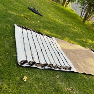 May include: A rectangular woven rug with a white, tan, and black striped pattern. The rug has a fringe of tassels along one edge. The rug is lying on a green grassy surface.