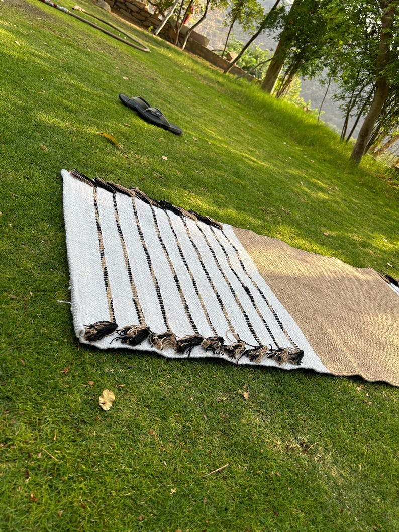 May include: A rectangular woven rug with a white, tan, and black striped pattern. The rug has a fringe of tassels along one edge. The rug is lying on a green grassy surface.