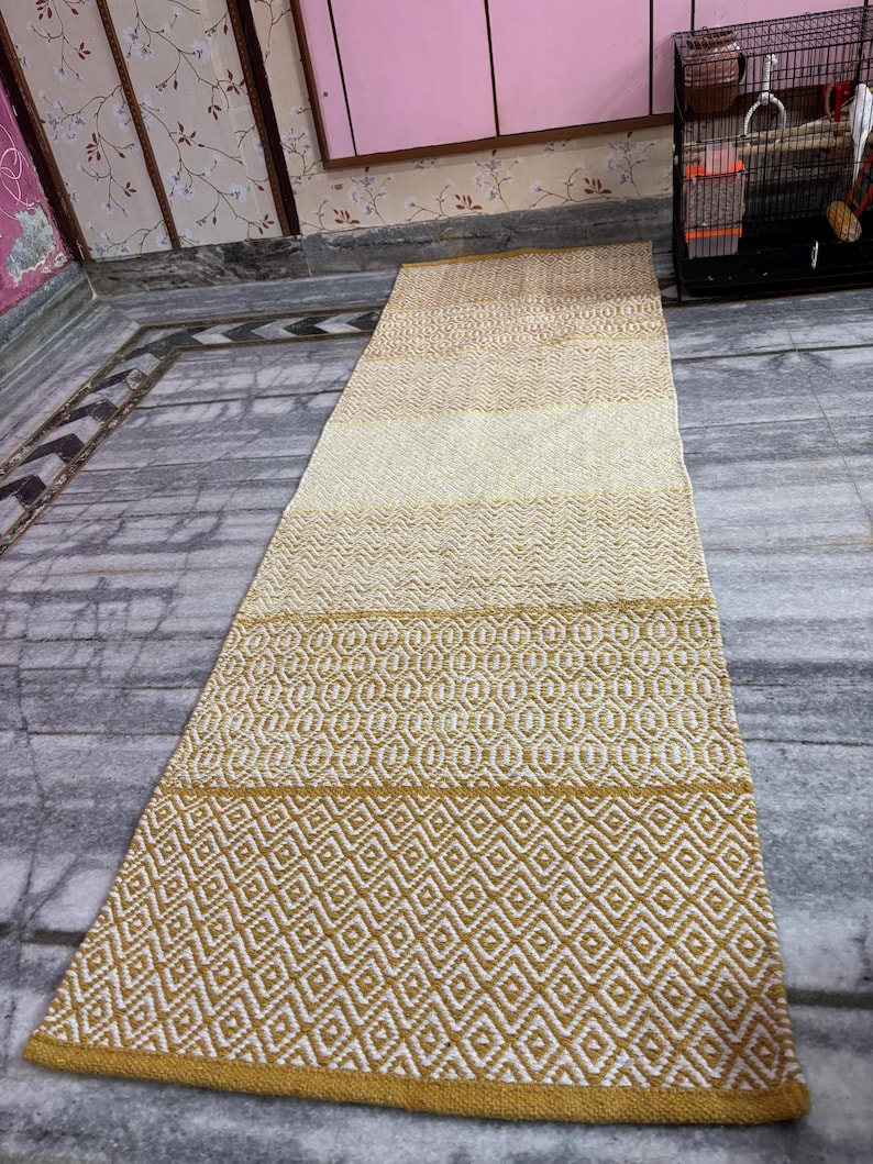 Puede incluir: Una alfombra larga y rectangular con un patr&oacute;n geom&eacute;trico. La alfombra presenta un degradado de colores, que va del beige claro al amarillo dorado. El dise&ntilde;o incluye formas de diamantes y &oacute;valos.