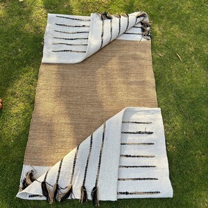 May include: A rectangular rug with a natural brown center and white ends. The white sections feature black and brown horizontal stripes and decorative tassels. The rug is partially folded, revealing the underside and is placed on green grass.