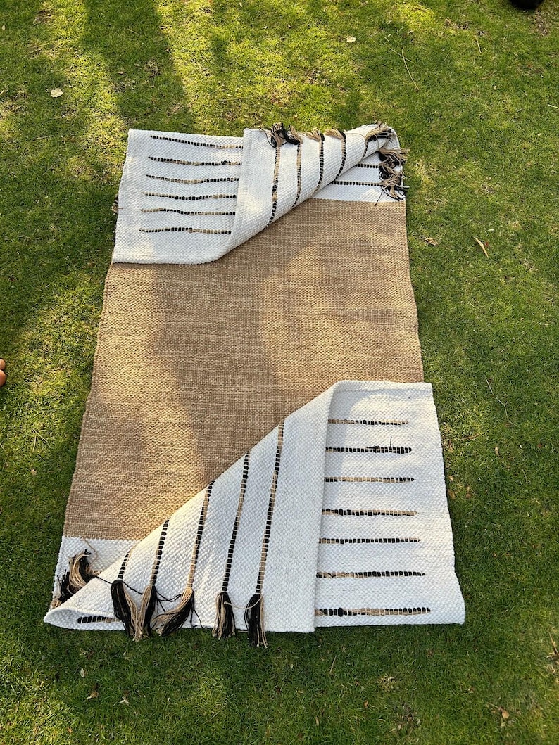 May include: A rectangular rug with a natural brown center and white ends. The white sections feature black and brown horizontal stripes and decorative tassels. The rug is partially folded, revealing the underside and is placed on green grass.