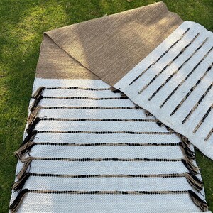 May include: Handwoven rug with a white base, accented by horizontal black and tan stripes. The rug features a tan border and decorative tassels. The design is simple and modern, suitable for various interior styles. The rug is placed on a green grassy surface.