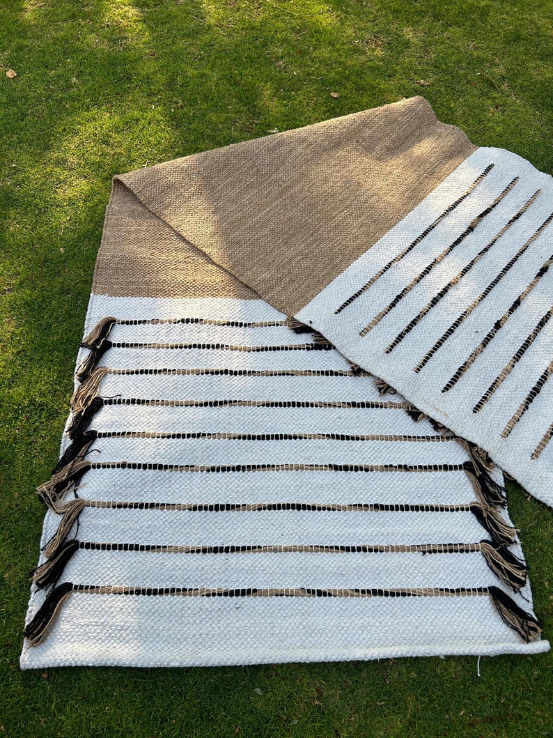 May include: Handwoven rug with a white base, accented by horizontal black and tan stripes. The rug features a tan border and decorative tassels. The design is simple and modern, suitable for various interior styles. The rug is placed on a green grassy surface.