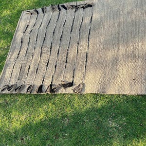 K&ouml;nnte beinhalten: Ein rechteckiger, gewebter Teppich in neutralem Beige mit vertikalen schwarzen Streifen. Der Teppich hat dekorative schwarze Quasten an einer Kante. Hergestellt aus Naturfaser, geeignet f&uuml;r den Innen- und Au&szlig;enbereich.