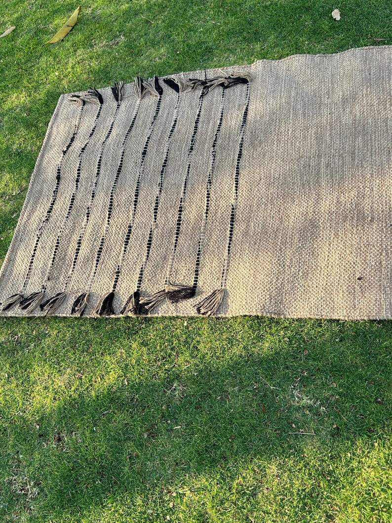 K&ouml;nnte beinhalten: Ein rechteckiger, gewebter Teppich in neutralem Beige mit vertikalen schwarzen Streifen. Der Teppich hat dekorative schwarze Quasten an einer Kante. Hergestellt aus Naturfaser, geeignet f&uuml;r den Innen- und Au&szlig;enbereich.