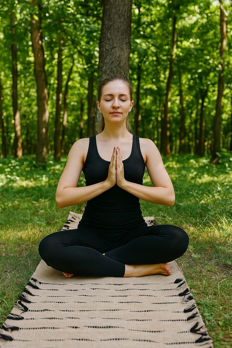 K&ouml;nnte beinhalten: Eine Person in schwarzer Kleidung sitzt im Schneidersitz auf einer beigen Yogamatte, die H&auml;nde in Gebetshaltung. Der Hintergrund ist ein gr&uuml;ner Wald mit hohen B&auml;umen, der eine ruhige Umgebung f&uuml;r Meditation und Wellness schafft.