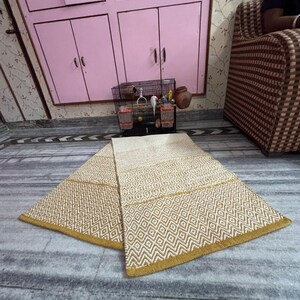 Puede incluir: Una alfombra beige y dorada con estampado geom&eacute;trico se muestra sobre un suelo de m&aacute;rmol. La alfombra presenta patrones de diamantes y zigzags con un borde dorado. La alfombra est&aacute; doblada por la mitad, mostrando su dise&ntilde;o y textura. El fondo incluye un armario rosa y un sof&aacute; estampado.