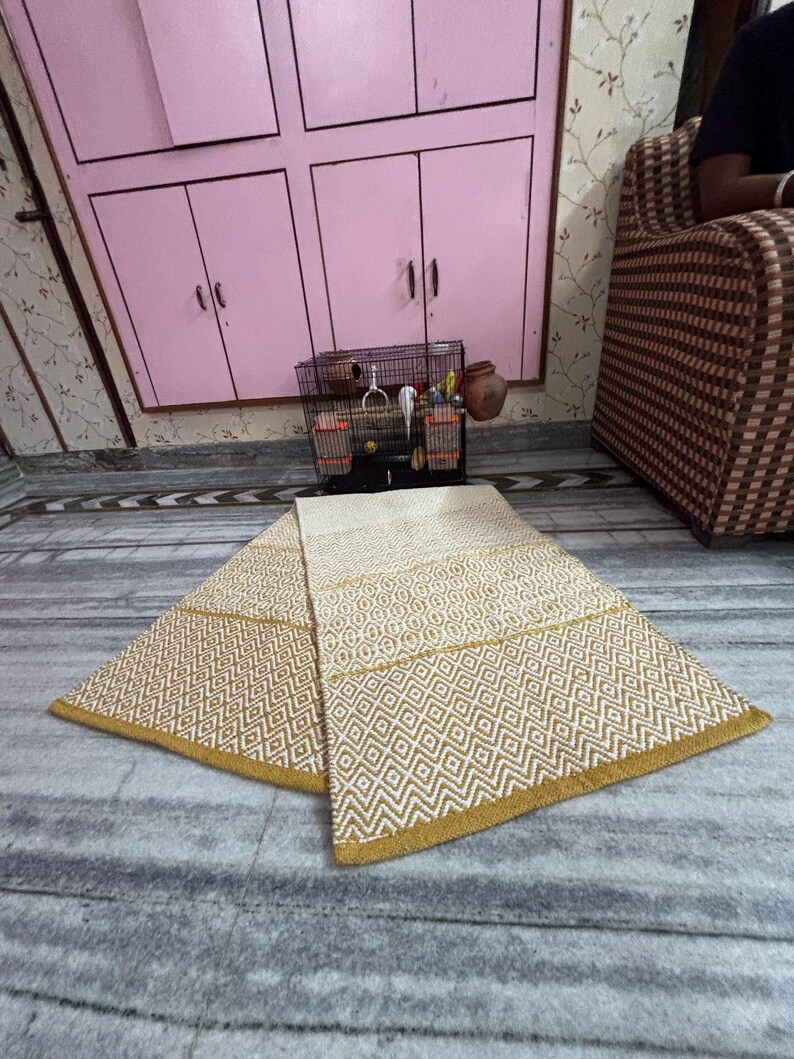 Puede incluir: Una alfombra beige y dorada con estampado geom&eacute;trico se muestra sobre un suelo de m&aacute;rmol. La alfombra presenta patrones de diamantes y zigzags con un borde dorado. La alfombra est&aacute; doblada por la mitad, mostrando su dise&ntilde;o y textura. El fondo incluye un armario rosa y un sof&aacute; estampado.