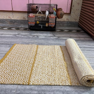 Puede incluir: Una alfombra rectangular enrollada, con un patr&oacute;n geom&eacute;trico de diamantes en crema y amarillo mostaza. La alfombra est&aacute; parcialmente desenrollada, revelando su textura y dise&ntilde;o. La alfombra est&aacute; hecha de un material tejido.