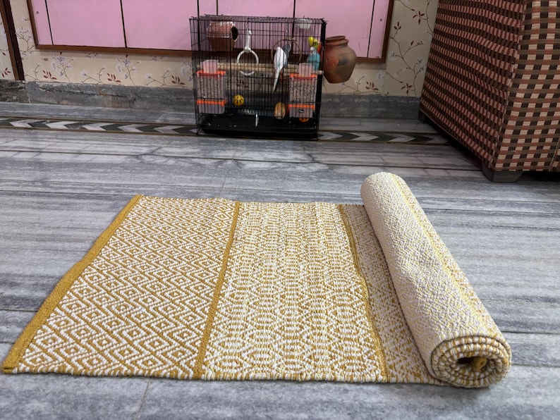 Puede incluir: Una alfombra rectangular enrollada, con un patr&oacute;n geom&eacute;trico de diamantes en crema y amarillo mostaza. La alfombra est&aacute; parcialmente desenrollada, revelando su textura y dise&ntilde;o. La alfombra est&aacute; hecha de un material tejido.