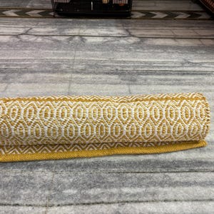 Puede incluir: Alfombra enrollada con un patr&oacute;n geom&eacute;trico amarillo y blanco. El dise&ntilde;o es una serie repetida de formas de diamante u ovaladas. La alfombra tiene un borde amarillo y est&aacute; hecha de material tejido. Est&aacute; sobre una superficie gris.