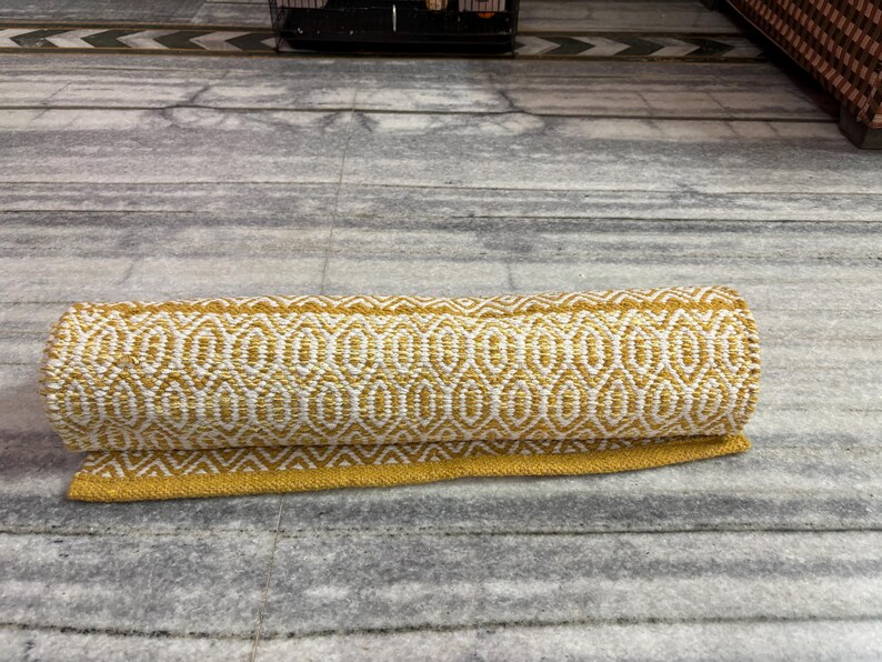 Puede incluir: Alfombra enrollada con un patr&oacute;n geom&eacute;trico amarillo y blanco. El dise&ntilde;o es una serie repetida de formas de diamante u ovaladas. La alfombra tiene un borde amarillo y est&aacute; hecha de material tejido. Est&aacute; sobre una superficie gris.