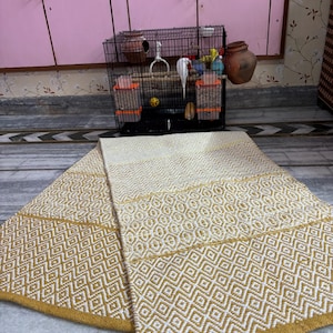 Puede incluir: Una alfombra plegada, de color crema, con un patr&oacute;n geom&eacute;trico de diamantes en dorado. La alfombra est&aacute; hecha de tela tejida y tiene un borde dorado. Una jaula para p&aacute;jaros con un p&aacute;jaro blanco y juguetes est&aacute; en el fondo.
