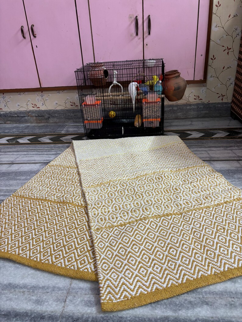 Puede incluir: Una alfombra plegada, de color crema, con un patr&oacute;n geom&eacute;trico de diamantes en dorado. La alfombra est&aacute; hecha de tela tejida y tiene un borde dorado. Una jaula para p&aacute;jaros con un p&aacute;jaro blanco y juguetes est&aacute; en el fondo.