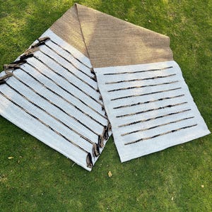 May include: A woven rug with a tan top section and a white bottom section with black and brown stripes. The rug has tassels on one end. The rug is lying on green grass.