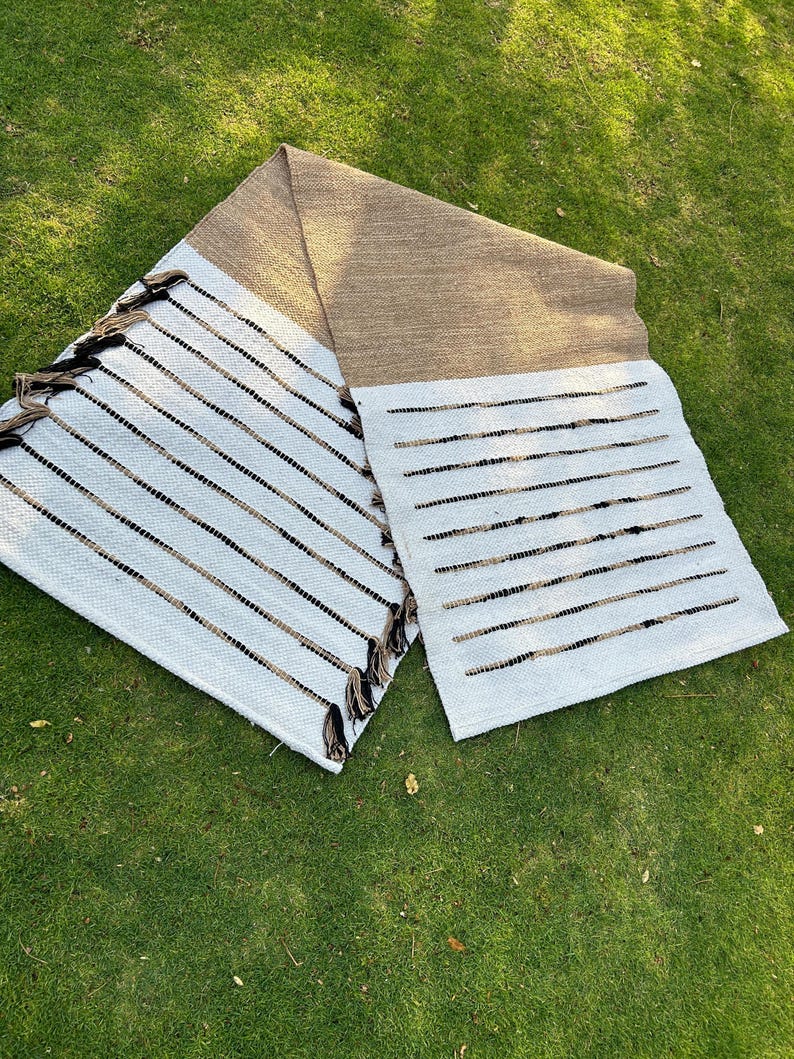 May include: A woven rug with a tan top section and a white bottom section with black and brown stripes. The rug has tassels on one end. The rug is lying on green grass.