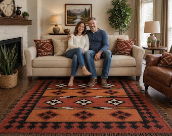May include: A living room scene featuring a couple seated on a beige sofa with patterned throw pillows. A vibrant, geometric patterned rug in shades of orange, black, and white covers the floor. A leather armchair and a fireplace complete the cozy setting.