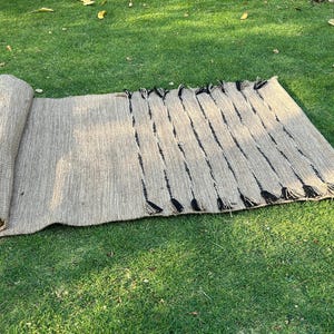 K&ouml;nnte beinhalten: Eine aufgerollte, rechteckige Matte aus Naturfasern, mit hellbrauner Farbe und schwarzen Akzenten. Die Matte ist teilweise auf einer gr&uuml;nen Grasfl&auml;che ausgerollt und zeigt ihre gewebte Textur und dekorative Fransen.