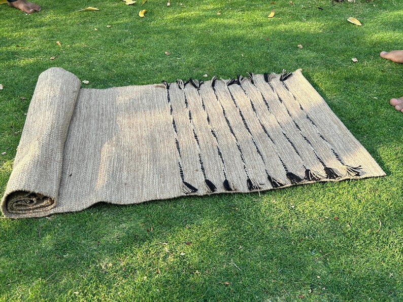 K&ouml;nnte beinhalten: Eine aufgerollte, rechteckige Matte aus Naturfasern, mit hellbrauner Farbe und schwarzen Akzenten. Die Matte ist teilweise auf einer gr&uuml;nen Grasfl&auml;che ausgerollt und zeigt ihre gewebte Textur und dekorative Fransen.