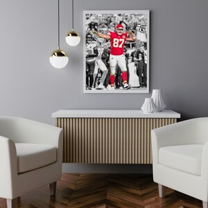 Puede incluir: Una impresión enmarcada de un jugador de fútbol americano con uniforme rojo, número 87, sobre un fondo en blanco y negro. El jugador celebra con los brazos levantados. La impresión está sobre un mueble de madera, flanqueado por dos sillones blancos.