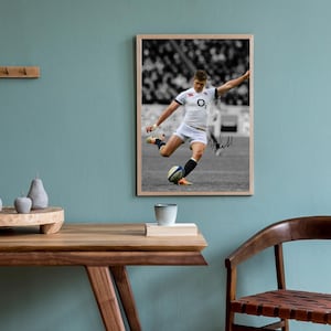Könnte beinhalten: Gerahmtes Foto eines Rugby-Spielers in Aktion, der einen Ball tritt. Der Spieler trägt ein weißes Trikot mit Logo und weiße Shorts. Der Hintergrund ist eine Schwarz-Weiß-Stadtszene. Der Rahmen hat eine helle Holzfarbe.