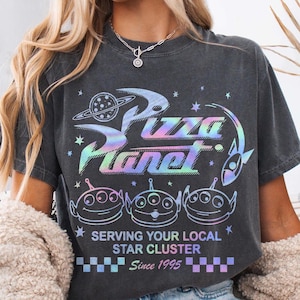 May include: Dark gray t-shirt with a holographic "Pizza Planet" graphic, featuring alien characters and the text "Serving Your Local Star Cluster Since 1995." The shirt has a crew neck and short sleeves.