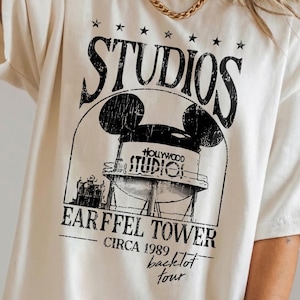 May include: Cream-colored t-shirt featuring a black graphic of the Hollywood Studios Earffel Tower with Mickey Mouse ears, the word "STUDIOS" above, and "EARFFEL TOWER CIRCA 1989 backlot tour" below.