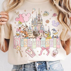 Puede incluir: Camiseta color crema con un colorido gr&aacute;fico de Disney World. El dise&ntilde;o incluye a Mickey y Minnie Mouse, el Pato Donald y otros personajes, junto con un castillo, palomitas de ma&iacute;z y la palabra "Disneyworld".