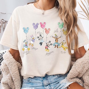Pode incluir: Camiseta creme com um gr&aacute;fico colorido de personagens da Disney segurando bal&otilde;es em forma de cabe&ccedil;a do Mickey Mouse. Os personagens incluem Pato Donald, Margarida, Mickey Mouse, Minnie Mouse, Pateta e Pluto. A camisa tem as assinaturas dos personagens.