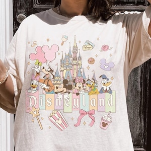 May include: Cream-colored t-shirt featuring a colorful Disneyland graphic. The design includes cartoon characters, a castle, and the word "Disneyland" in pastel colors. Additional elements include popcorn, a bow, and a drink.