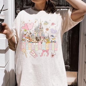 Puede incluir: Camiseta color crema con un gr&aacute;fico colorido de personajes de Disney y un castillo. La palabra "Disneyworld" est&aacute; impresa en letras pastel en la parte inferior. Otros gr&aacute;ficos incluyen una caja de palomitas de ma&iacute;z, un lazo y una bebida.