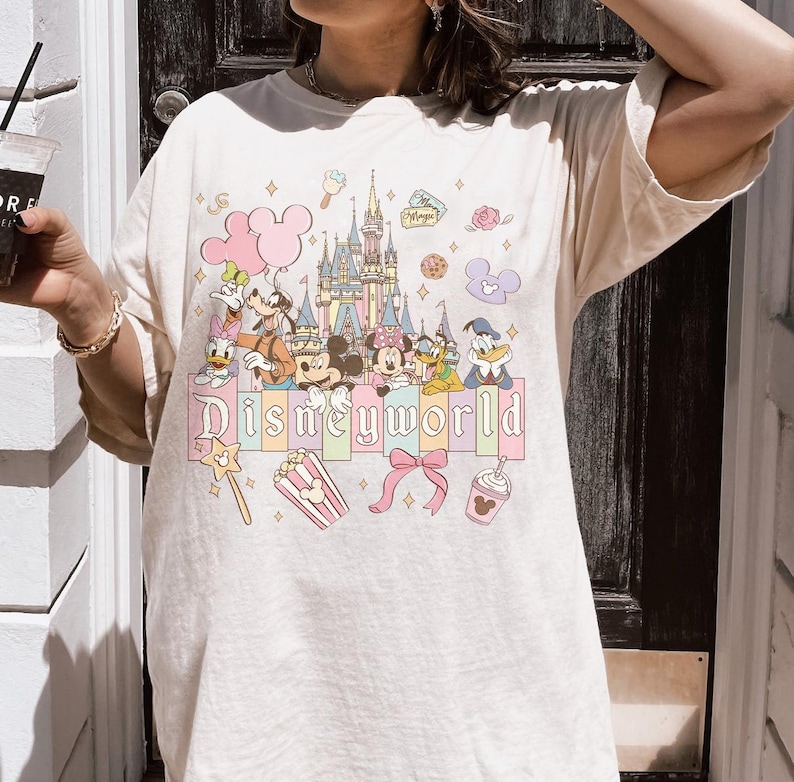 Puede incluir: Camiseta color crema con un gr&aacute;fico colorido de personajes de Disney y un castillo. La palabra "Disneyworld" est&aacute; impresa en letras pastel en la parte inferior. Otros gr&aacute;ficos incluyen una caja de palomitas de ma&iacute;z, un lazo y una bebida.