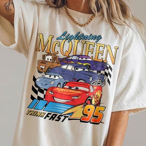 Puede incluir: Camiseta de color crema con un gr&aacute;fico de Rayo McQueen y otros personajes de la pel&iacute;cula Cars. El dise&ntilde;o incluye el texto "Lightning McQueen", "Think Fast" y el n&uacute;mero "95".