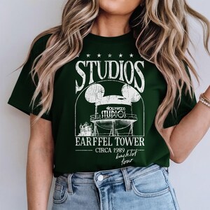 May include: Dark green t-shirt with a white graphic featuring the word "STUDIOS" above a Mickey Mouse-shaped water tower design. The text "EARFFEL TOWER CIRCA 1989 backlot tour" is below the design.