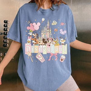 May include: A light blue t-shirt featuring a colorful Disneyland graphic. The design includes Mickey Mouse, Minnie Mouse, Donald Duck, and other Disney characters, along with the Disneyland castle and the word "Disneyland".