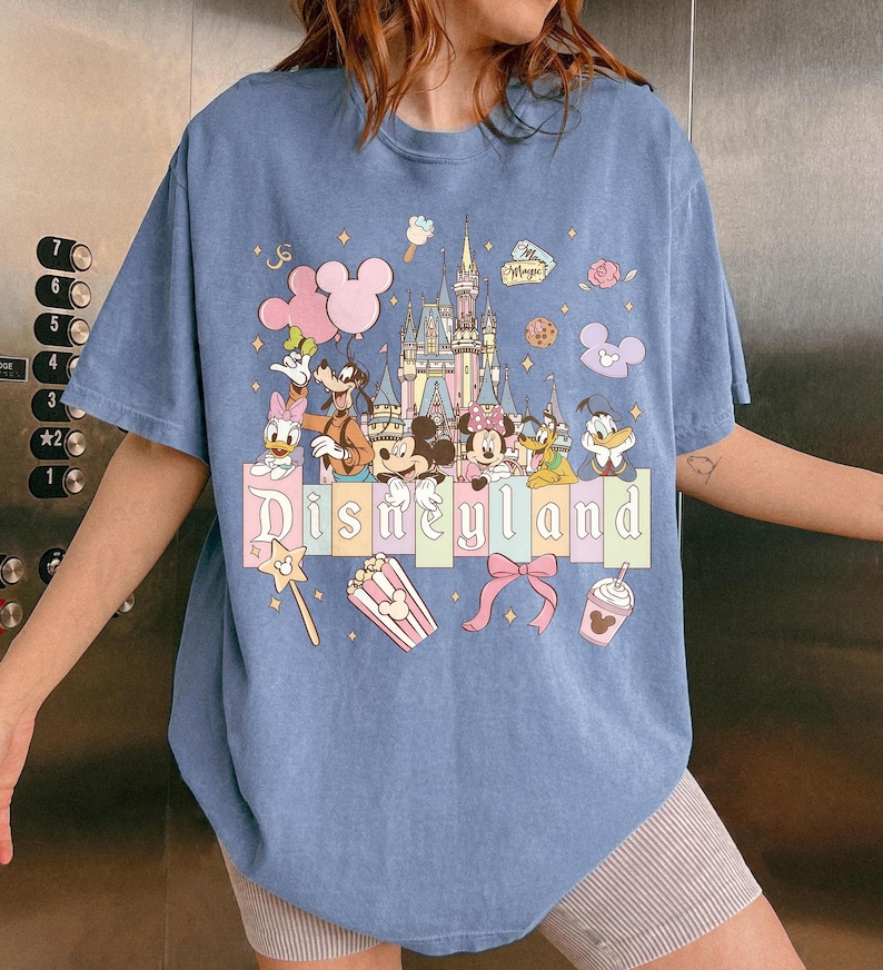 May include: A light blue t-shirt featuring a colorful Disneyland graphic. The design includes Mickey Mouse, Minnie Mouse, Donald Duck, and other Disney characters, along with the Disneyland castle and the word "Disneyland".