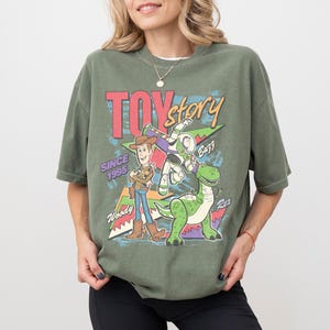 Op de afbeelding: Olijfgroen T-shirt met de woorden "Toy Story" in rood en geel, met Woody, Buzz Lightyear en Rex de dinosaurus. Het shirt heeft ook de tekst "Since 1995".