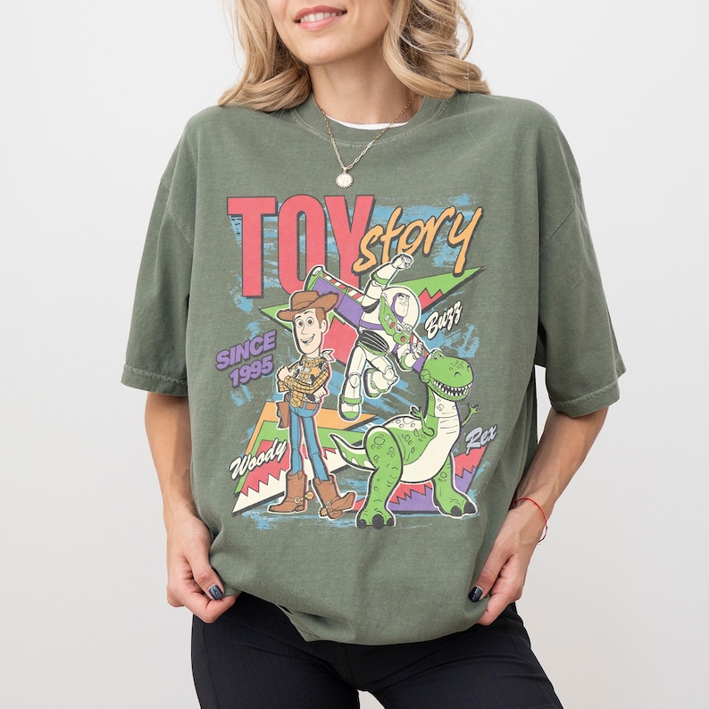 Op de afbeelding: Olijfgroen T-shirt met de woorden "Toy Story" in rood en geel, met Woody, Buzz Lightyear en Rex de dinosaurus. Het shirt heeft ook de tekst "Since 1995".