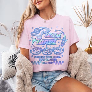 May include: A light pink t-shirt featuring the Pizza Planet logo in iridescent blue and green. Below the logo are three alien characters. The text "Serving Your Local Star Cluster" and "Since 1995" are also printed on the shirt.