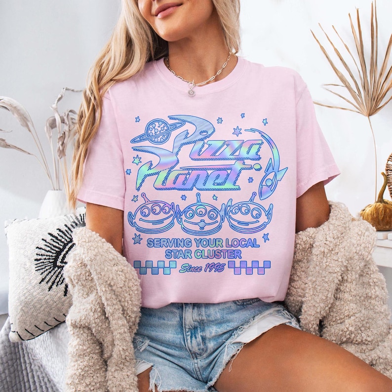 May include: A light pink t-shirt featuring the Pizza Planet logo in iridescent blue and green. Below the logo are three alien characters. The text "Serving Your Local Star Cluster" and "Since 1995" are also printed on the shirt.