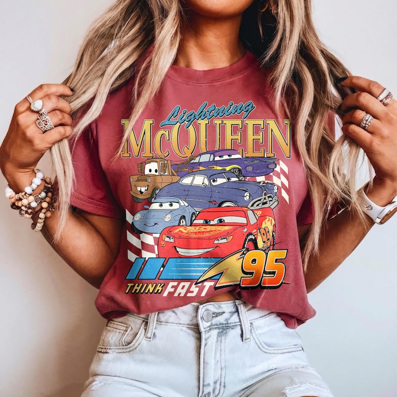 Puede incluir: Una camiseta granate con un dise&ntilde;o de Rayo McQueen y otros personajes de Cars. El dise&ntilde;o incluye el texto "Lightning McQueen" y "95", con la frase "Think Fast" debajo. La camiseta tiene un aspecto vintage y desgastado.